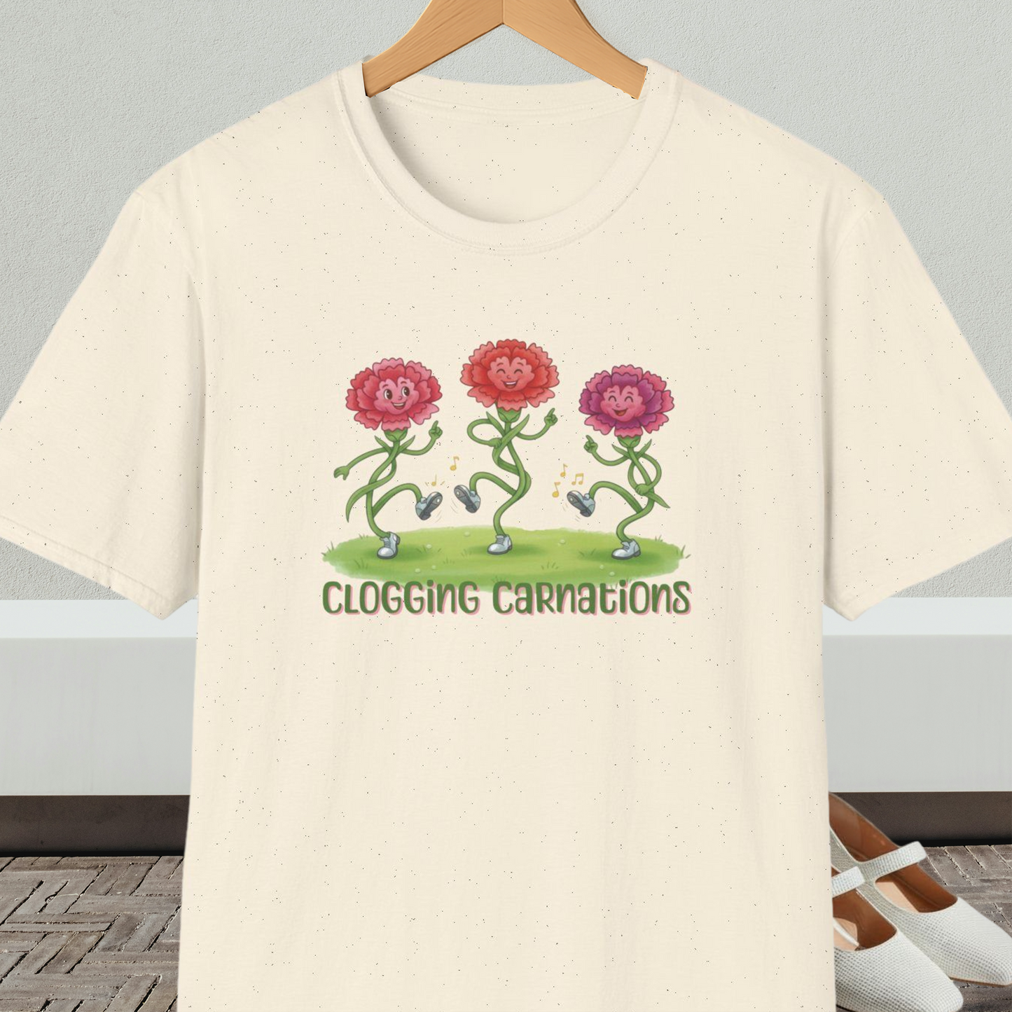 Natural beige t-shirt with dancing flower design and text 'Clogging Carnations', on a hanger in front of a neutral background.
